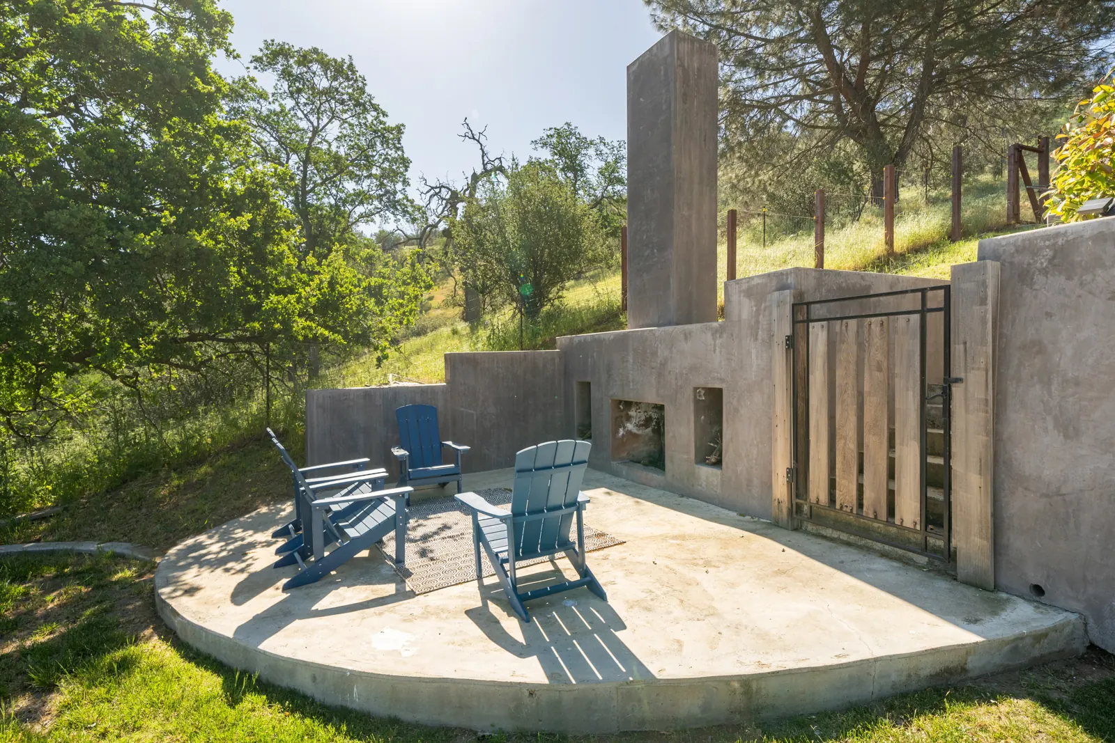 Round concrete patio with four blue Adirondack chairs facing the outdoor fireplace, among oak trees